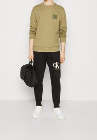 Felpa verde oliva con una piccola tasca con logo, abbinata a pantaloni jogger neri con un grande logo. Sneakers bianche e una borsa nera completano il look.