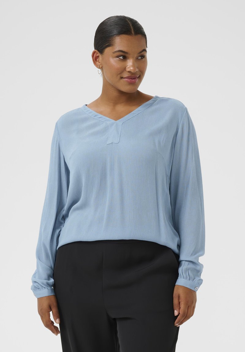 Haut à manches longues bleu clair avec un col en V, présentant une texture légèrement rayée et des accents de boutons sur les manches. Assorti avec un pantalon noir.