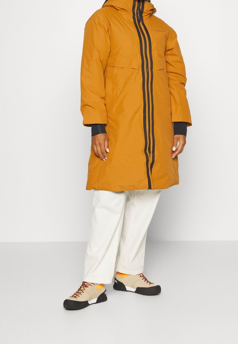 Brun lang frakke med hætte, sort lodret lynlåsdetaile, og elastiske manchetter, parret med cremefarvede bukser og beige-orange vandresko.