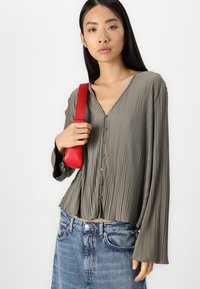 Olivgrünes, plissiertes Blusenshirt mit langen Ärmeln und V-Ausschnitt, mit Knöpfen vorne. Kombiniert mit blauem Jeansrock und roter Handtasche.