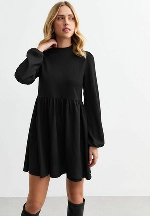 Mujer joven con cabello rubio, vestida con un vestido negro de manga larga y botas negras, de pie contra un fondo blanco liso.