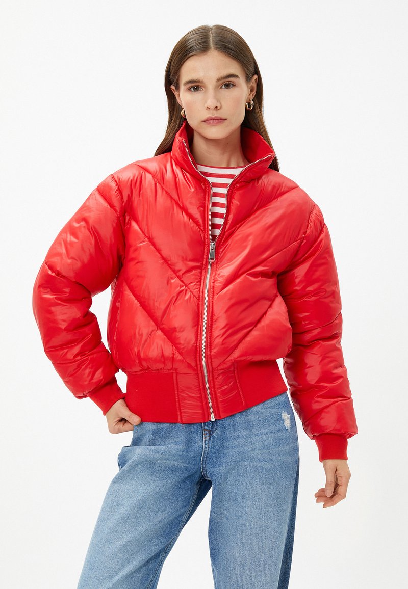 Rød dunjakke med høj krave, diagonal quiltet mønster, lynlås og ribbede manchetter. Båret over en stribet bluse og parret med blå jeans.