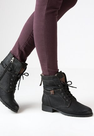 Persona con botines negros de tacón bajo con cordones y cremalleras laterales, y pantalones ajustados color burdeos, de pie con un pie ligeramente levantado.