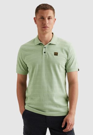 Mann trägt hellgrünes, strukturiertes Poloshirt mit kurzen Ärmeln und schwarze Hose, steht mit einer Hand in der Tasche.