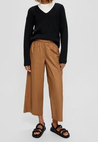 Zwarte gebreide trui met V-hals, gecombineerd met losse, cropped bruine broeken en zwarte sandalen met bandjes. Zachte textuur, casual stijl.