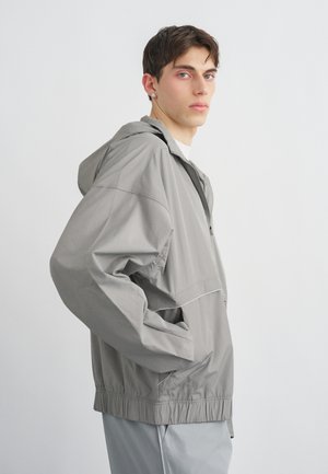 Jonge man met kort bruin haar die een lichtgrijs windjack met capuchon draagt, met zijn handen in de zakken, lichtjes zijwaarts gericht.