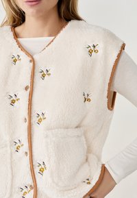 Gilet in pile senza maniche di colore crema con bordi marroni, caratterizzato da ricami floreali e due tasche frontali. Texture morbida e chiusura a bottoni.