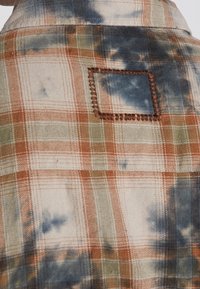 Tissu à carreaux avec des carrés orange, beige et verts tachés de teinture bleue et une petite pièce rectangulaire cousue avec un fil brun.