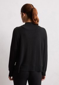 Donna con un ponytail castano-rossiccio, che indossa un maglione nero a maniche lunghe e pantaloni neri, rivolta verso un muro bianco e testurizzato, vista da dietro.
