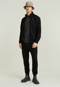 Chaqueta negra de poliéster con cremallera sobre una camiseta oscura, combinada con pantalones jogger negros y botas de goma negras. Un sombrero de cubo oliva completa el atuendo.