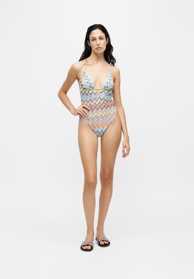 Femme portant un maillot de bain une pièce à motif zigzag coloré et des sandales assorties, debout devant un fond gris uni.