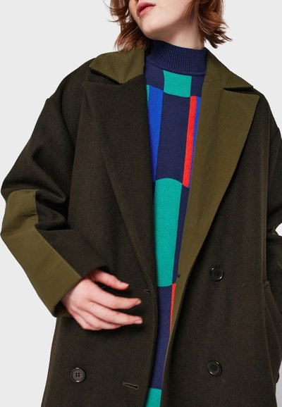 Olive green wool coat with wide lapels, textured fabric, double-button closure, and large pockets, worn over a multicolored patterned sweater.