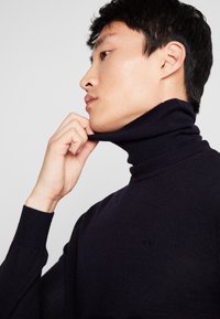Marineblå turtleneck sweater i blødt strikmateriale med en tætsiddende pasform og ribstrikkede manchetter. Nærbillede af halsområdet, der justeres.