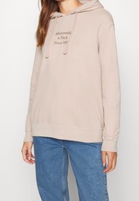 Sudadera beige con capucha ajustable, logo bordado en marrón oscuro y detalles de costura visibles. Combinada con jeans azules.