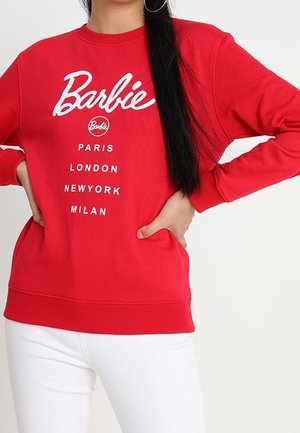 Personne portant un sweat-shirt Barbie rouge avec les noms des villes Paris, Londres, New York, Milan, associé à un pantalon blanc.