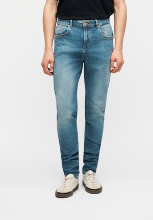 Person wearing light blue slim-fit jeans, black top, and white sneakers, standing against a plain light background.