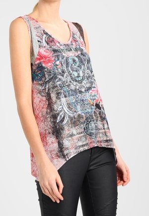 Kobieta w różowym, asymetrycznym topie na ramiączkach z misternymi czarnymi, niebieskimi i czerwonymi wzorami kwiatowymi i abstrakcyjnymi, w połączeniu z czarnymi spodniami.