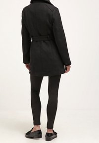 Henkilö, jolla on päällään musta vyöllinen takki, mustat kapeat housut ja mustat slip-on-kengät, seisoo selkä menosuuntaan yksinkertaista taustaa vasten.