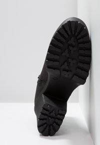 Semelle de bottine noire avec un motif de bande de roulement profond et robuste, positionnée verticalement contre un fond de sol et de mur blancs.