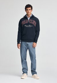 Marinblå sweatshirt med "POLO CLUB"-logotyp, halvdragkedja och ribbad nederkant, matchad med ljusblå jeans och beige sneakers.