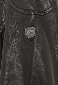 Dettaglio di una giacca in pelle nera con superficie lucida, finitura testurizzata e un emblema metallico con la parola "Gipsy" su uno scudo.