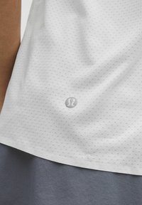 Wit sportshirt van een textuurstof met een patroon, voorzien van een grijs logo in het midden onderaan. De zoom heeft een lichte kromming.