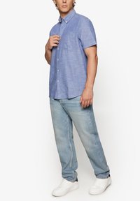 Camisa de manga corta azul claro con cuello abotonado, combinada con jeans de mezclilla de lavado claro y zapatillas blancas. Presenta un detalle de bolsillo.