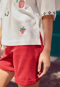 Vit kortärmad skjorta med färgglad fruktbroderi och blå accenter, parat med knälånga röda shorts med upprullade fållar.