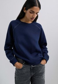 Marineblaues Sweatshirt mit Rundhalsausschnitt und Raglanärmeln; mit kontrastierenden Nähten und lockerer Passform; kombiniert mit dunkelgrauen Jeans.