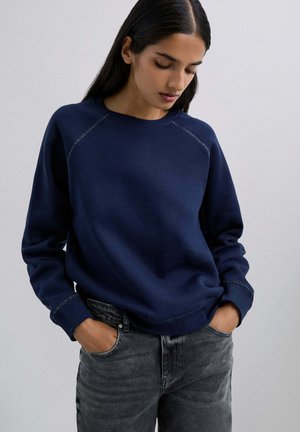 Sudadera azul marino con cuello redondo y mangas raglán; presenta costuras en contraste y un ajuste relajado; combinada con jeans gris oscuro.