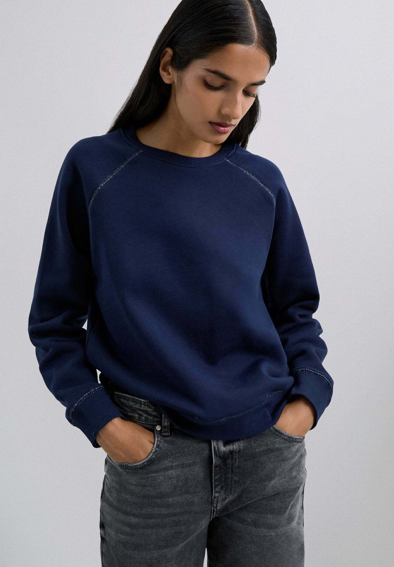 Marineblaues Sweatshirt mit Rundhalsausschnitt und Raglanärmeln; mit kontrastierenden Nähten und lockerer Passform; kombiniert mit dunkelgrauen Jeans.
