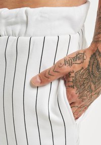 Pantalons en tissu blanc avec des rayures verticales noires, dotés d'une large ceinture élastique et de poches latérales. Un doigt est visible dans la poche.