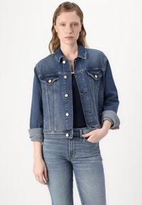 Denim jacket in medium blue with long sleeves, buttoned front, two chest pockets, and rolled cuffs. Worn over a dark top, paired with jeans.