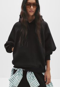 Hoodie noir surdimensionné avec une finition texturée, doté d'une capuche à cordon. Une chemise rayée verte et blanche est nouée à la taille.