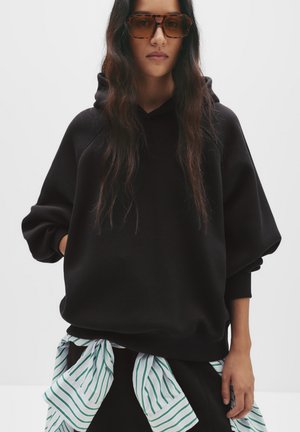 Hoodie noir surdimensionné avec une finition texturée, doté d'une capuche à cordon. Une chemise rayée verte et blanche est nouée à la taille.