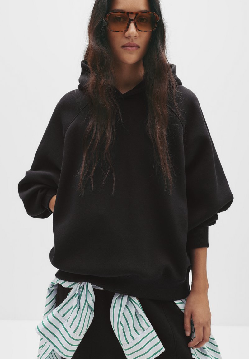 Hoodie noir surdimensionné avec une finition texturée, doté d'une capuche à cordon. Une chemise rayée verte et blanche est nouée à la taille.