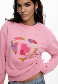 Rožinis džemperis su širdies dizainu ir daugiakoliažinėmis siuvinėjimo detalėmis. Pagamintas iš minkštos medžiagos, su apvaliomis iškirptėmis ir ribinėmis rankogalais.