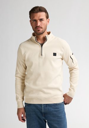 Maglione beige con zip e colletto alto, maniche lunghe, texture morbida e un piccolo logo nero sul petto. Abbinato a jeans in denim blu.