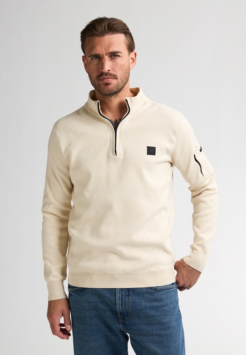 Maglione beige con zip e colletto alto, maniche lunghe, texture morbida e un piccolo logo nero sul petto. Abbinato a jeans in denim blu.