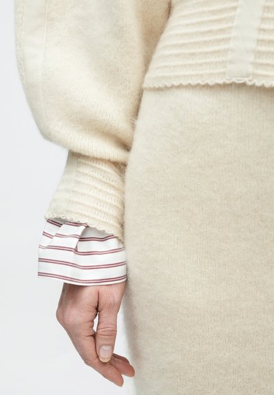 Beežikas kudumiga kampsun, millel on sooniktekstuur ja lainelise äärega serv, kantud üle valge särgi, millel on kitsad burgundiapunased triibud, näidatud käega.