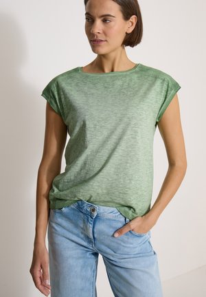Femme aux cheveux courts bruns portant un haut sans manches vert et un jean bleu clair, une main dans la poche, sur fond uni.