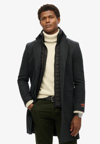 Ciemnoszary płaszcz na czarnej kurtce puchowej, o dopasowanym kroju, z guzikami oraz z grubym kremowym golfem pod spodem.