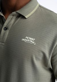 Olivgrön pikétröja med texturerad yta, utrustad med en gul randig krage och en vit logotyp som säger "PETROL INDUSTRIES."