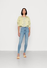 Jaqueta cropped verde claro sobre uma blusa floral, combinada com jeans de cintura alta azul claro e sandálias amarelas tipo slip-on. Fundo simples.