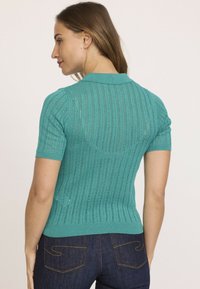 Pull à manches courtes, ajusté, de couleur sarcelle, présentant un motif tricoté texturé ainsi qu'un col et un ourlet à côtes. La vue arrière révèle une silhouette lisse.