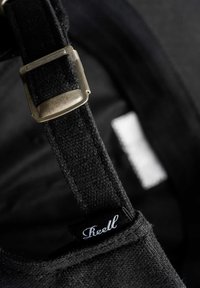 Schwarzes Stoffband mit einer metallischen Schnalle, die eine strukturierte Oberfläche aufweist und mit einem kleinen schwarzen Etikett mit weißem Text versehen ist. Minimalistisches Design mit Kontrasten.