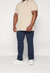 Beżowa koszulka polo z czarnym logo, kontrastującym kołnierzykiem, w połączeniu z granatowymi spodniami i białymi trampkami. Tkanina wydaje się gładka i casualowa.