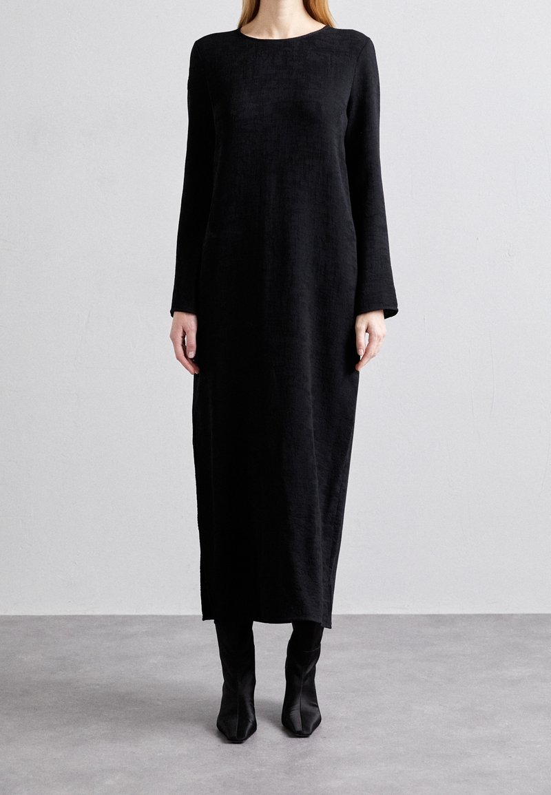 Femme portant une longue robe noire texturée à manches longues et des bottes noires pointues, debout contre un mur gris clair.