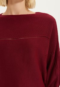 Pull en maille bordeaux à coupe ample, présentant un motif texturé et un large décolleté. Élément de design horizontal notable au-dessus de la poitrine.