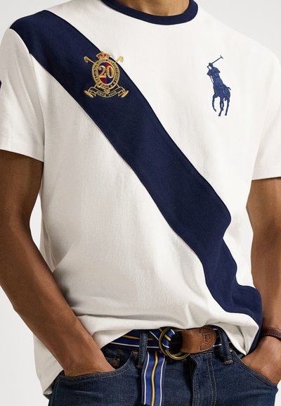 Λευκό T-shirt με κοντά μανίκια και σκούρα μπλε λοξή ρίγα, κεντημένο έμβλημα με τον αριθμό "20" και μικρό λογότυπο με παίκτη πόλο. Συνδυάζεται με τζιν παντελόνι.